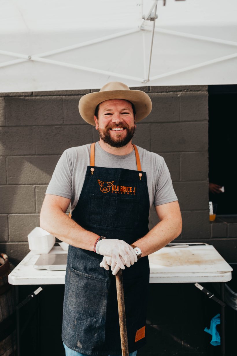 Smiling bearded man in a hat and denim Old Bruce J Barbeque apron leaning on a tool at an outdoor barbecue stall.